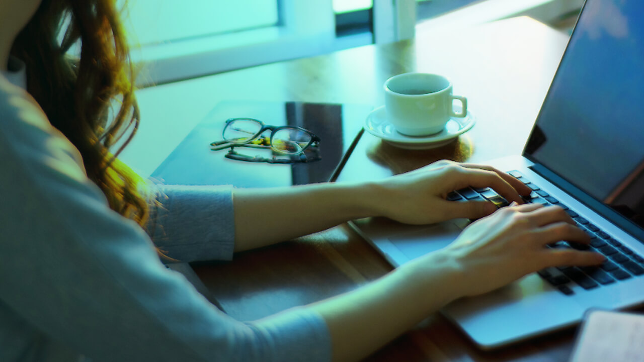 Woman typing on her laptop with a coffee cup, glasses and notebook next to her